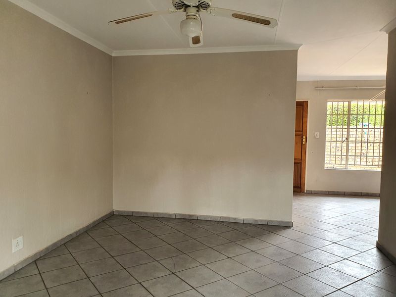 An empty, open-plan living area featuring neutral tiled flooring, a ceiling fan, and natural light through a window with security bars.