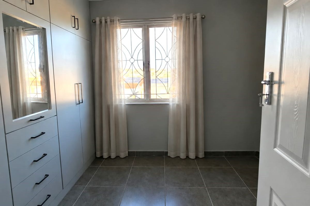 A clean, empty bedroom featuring modern built-in cupboards with integrated mirror, tiled flooring, and a window fitted with a security gate.