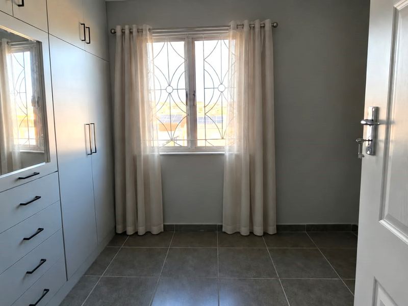 A clean, empty bedroom featuring modern built-in cupboards with integrated mirror, tiled flooring, and a window fitted with a security gate.