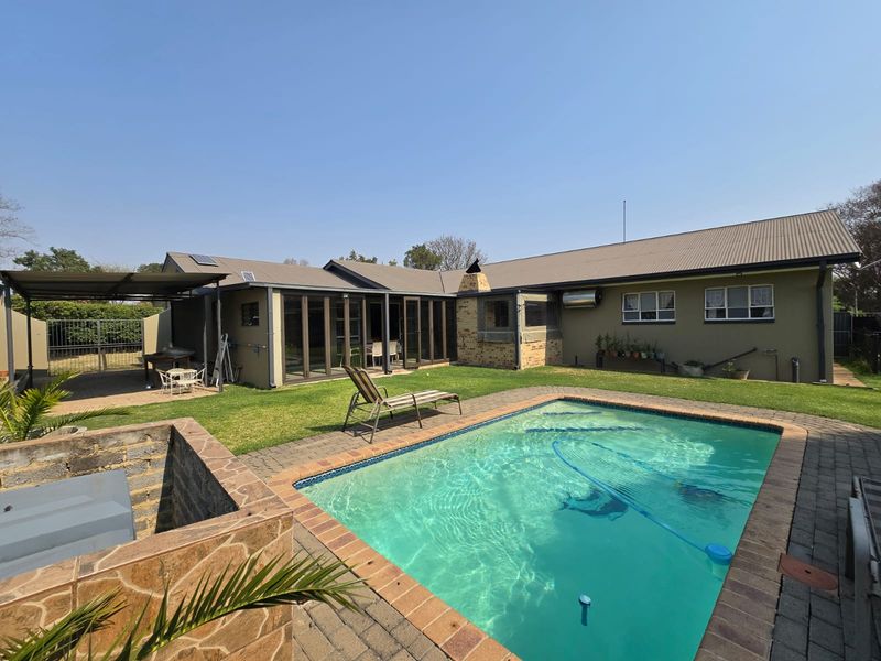 Rear exterior view featuring a swimming pool, paved surround, covered patio area, and a built-in brick braai against the house facade.