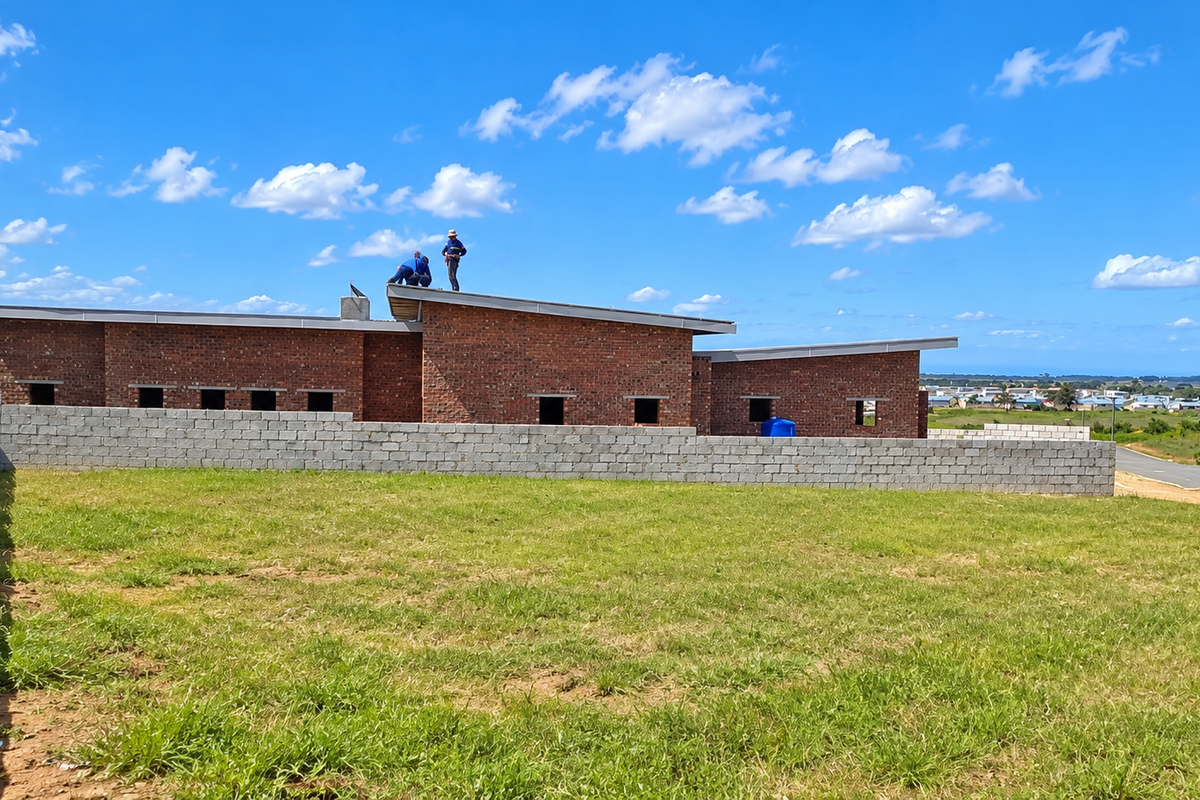 A rear view of a house under construction, featuring exposed brick walls, a mono-pitch roof, and a concrete block boundary wall on a grassy plot.