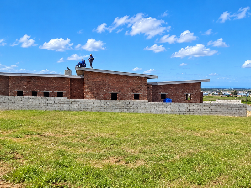 A rear view of a house under construction, featuring exposed brick walls, a mono-pitch roof, and a concrete block boundary wall on a grassy plot.