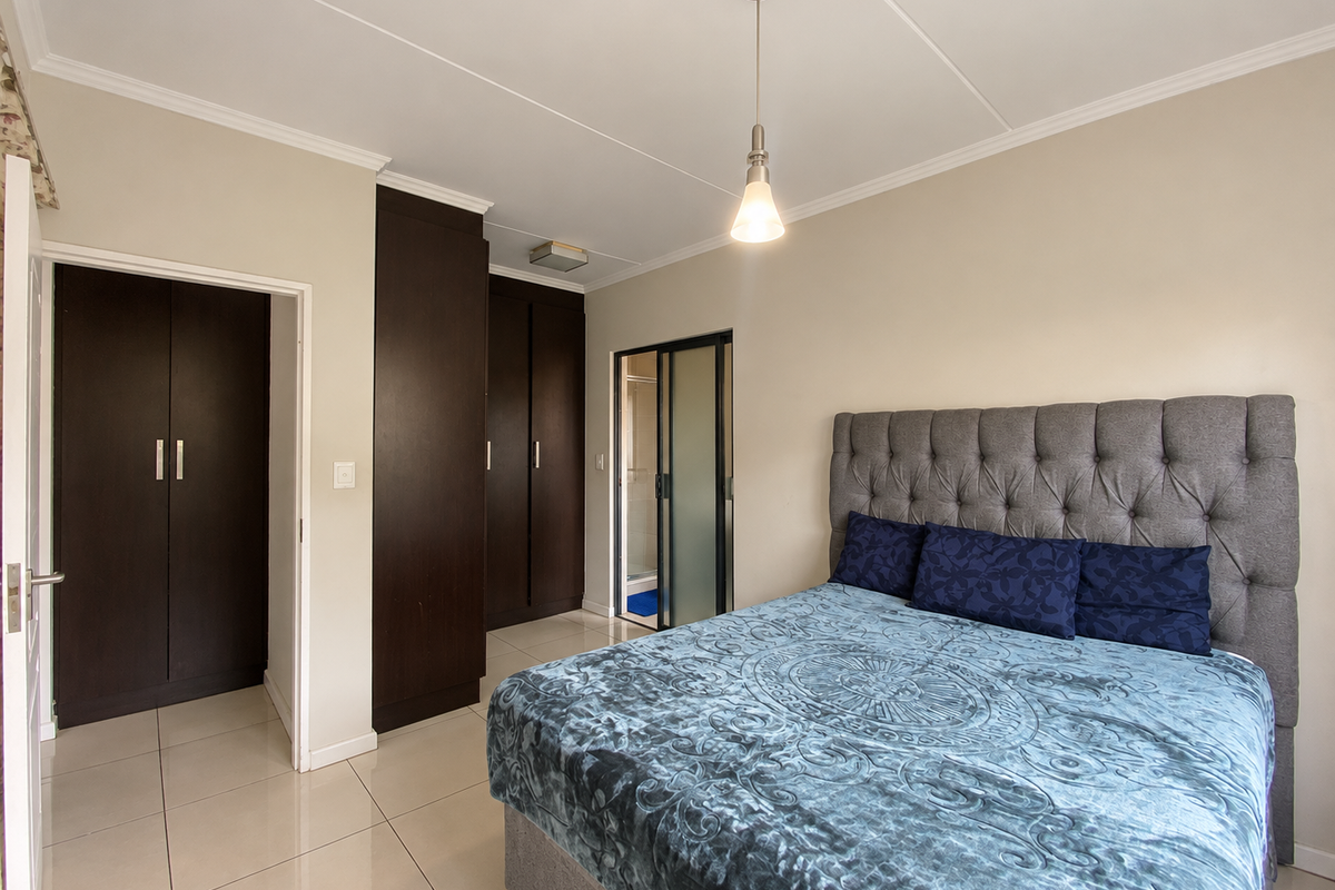 A well-maintained bedroom featuring dark wood built-in cupboards, neutral tiled flooring, and an en-suite bathroom access via a sliding door.