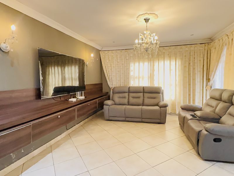 A spacious living room featuring tiled flooring, a large built-in media unit, decorative ceiling rose with chandelier, and elegant wall sconces.