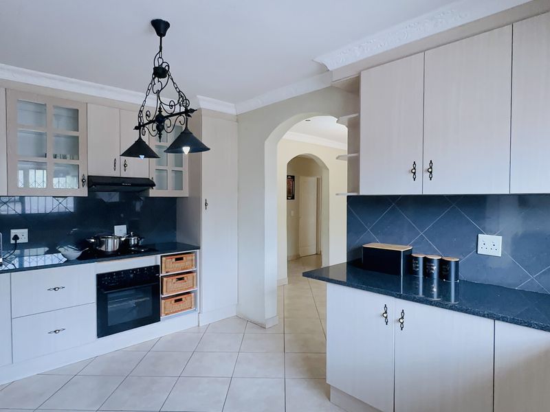 A well-maintained kitchen featuring light-toned cabinetry, dark granite countertops, built-in oven and hob, and tiled flooring, leading into an arched hallway.