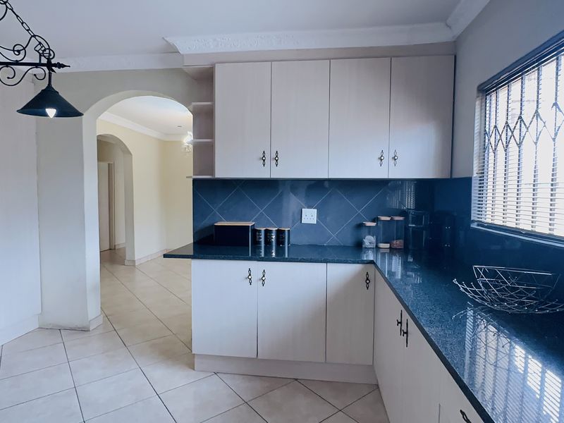 A functional kitchen featuring light-toned cabinetry, dark granite countertops, tiled backsplash, and ceramic flooring, with an open archway leading to adjacent living areas.