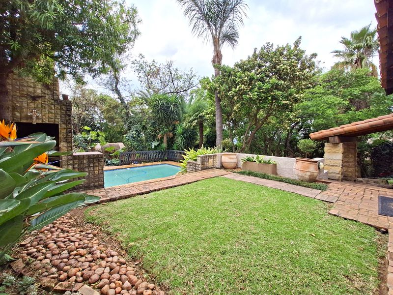 A private outdoor pool area featuring a swimming pool, brick-paved patio, and a built-in braai, surrounded by established garden landscaping.