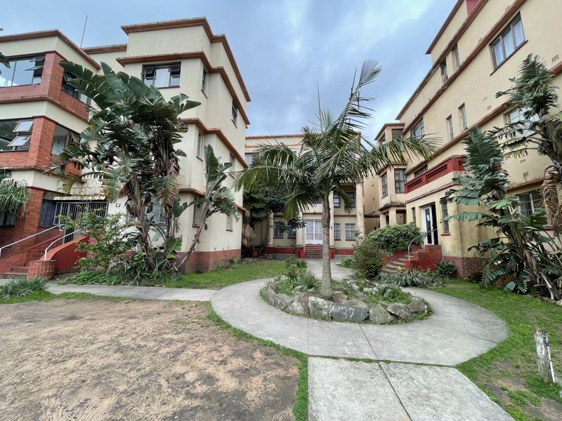 Exterior view of an apartment complex courtyard featuring concrete pathways, established garden beds, and multi-story building facades with visible wear.