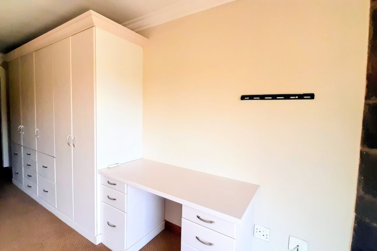 A bedroom featuring a large built-in wardrobe and integrated desk unit, with neutral walls and carpet flooring.