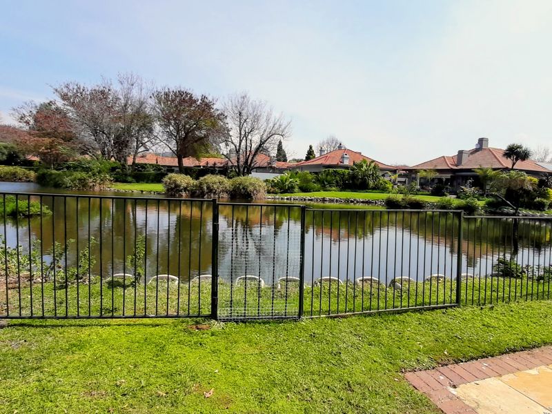 A scenic view of a tranquil pond surrounded by landscaped gardens and residential properties, featuring a secure metal perimeter fence.