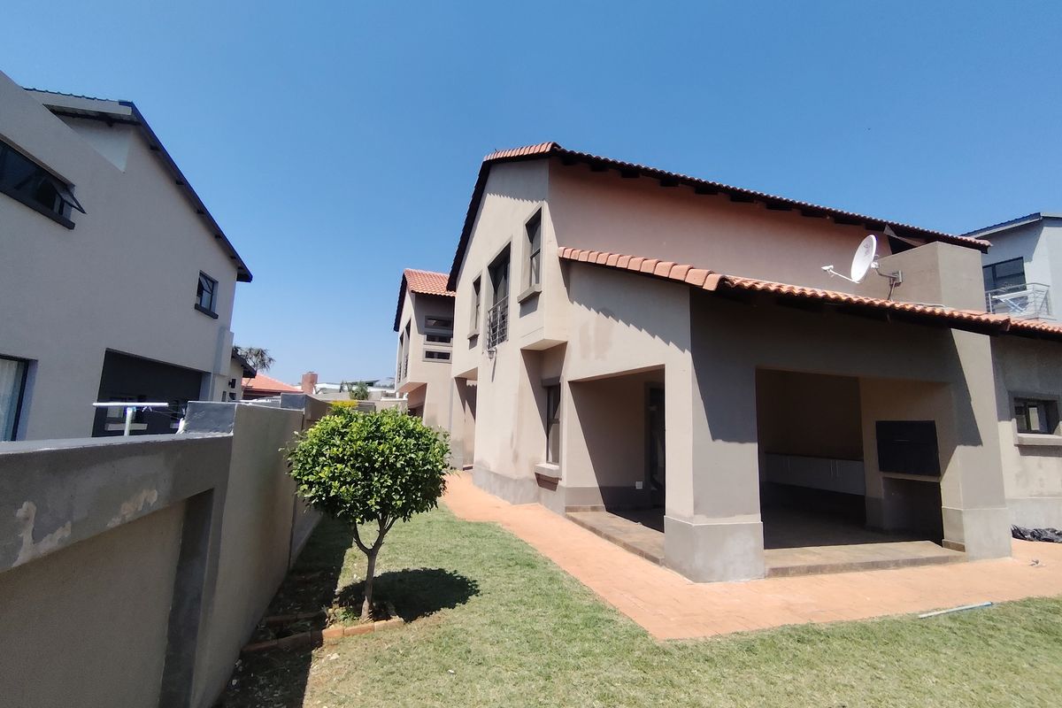 Rear view of the property featuring a covered patio with a built-in braai, a small lawn, and a paved walkway along the boundary wall.