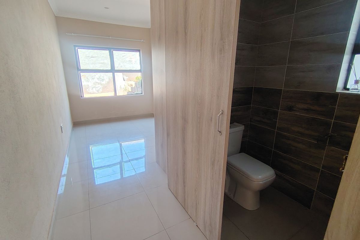 A bright, empty bedroom featuring modern tiled flooring and an en-suite bathroom area with a sliding door and dark feature wall tiling.