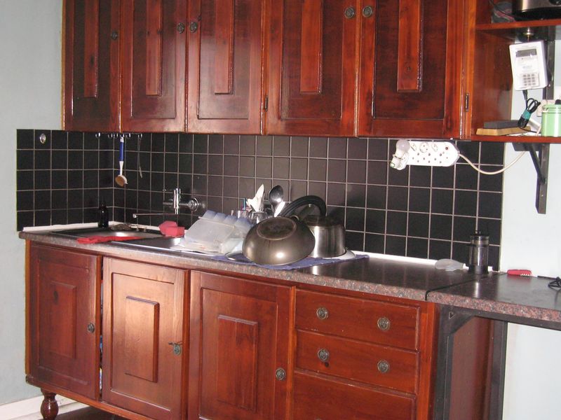 A dated kitchen area featuring dark wooden cabinetry, a black tiled backsplash, and a functional stainless steel sink with wall-mounted hardware.
