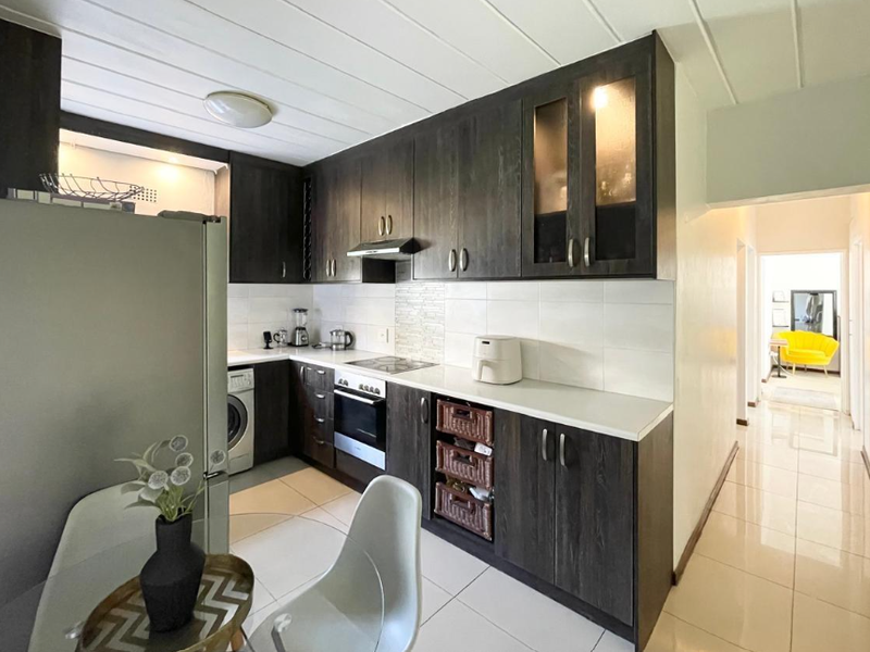 A functional kitchen featuring dark wood cabinetry, white countertops, tiled backsplash, and integrated appliances, with direct access to the hallway.