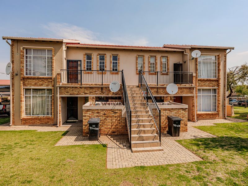 Rear view of a two-story apartment building featuring brick facade, external staircase, paved walkways, and communal garden space.