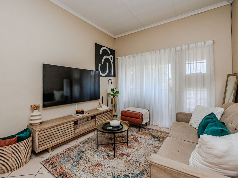 A cozy living room featuring tiled flooring and large windows with sheer curtains, providing natural light and access to the exterior.