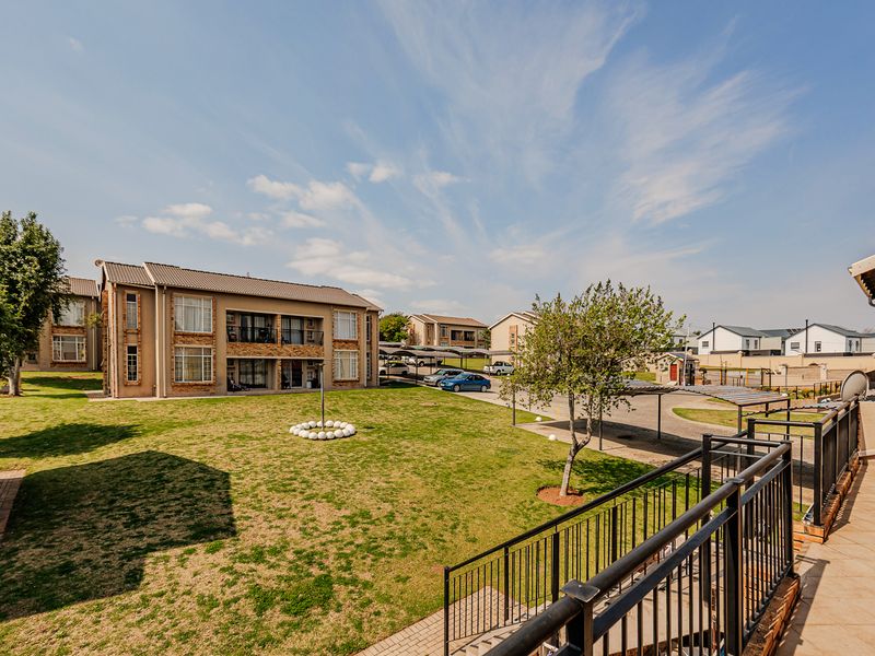 A view from a balcony overlooking a communal garden area, paved driveway, and apartment complex buildings with carports.