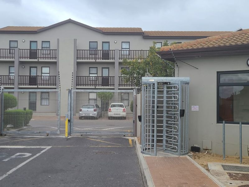Exterior view of an apartment complex entrance featuring a security gate, turnstile, guard house, and paved driveway with electric fencing.