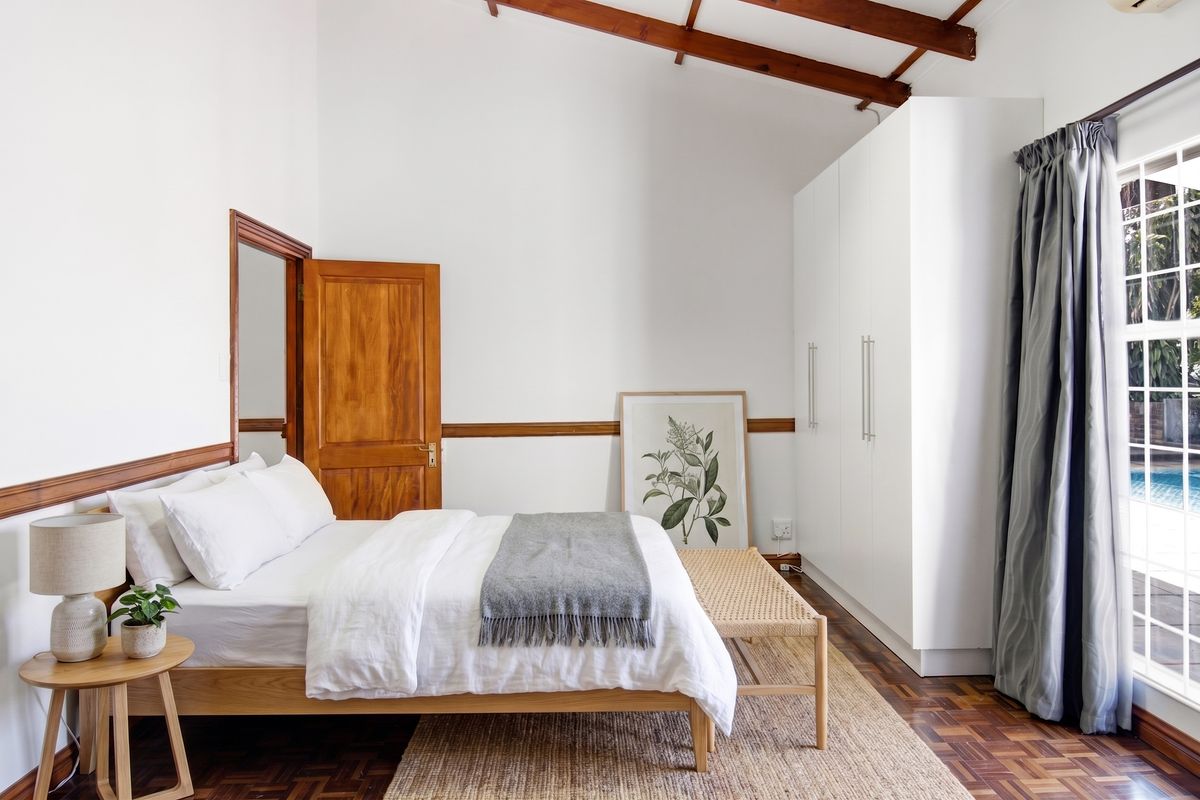 Bright bedroom featuring exposed timber ceiling beams, parquet flooring, built-in wardrobes, and a sliding glass door providing access to the pool area.