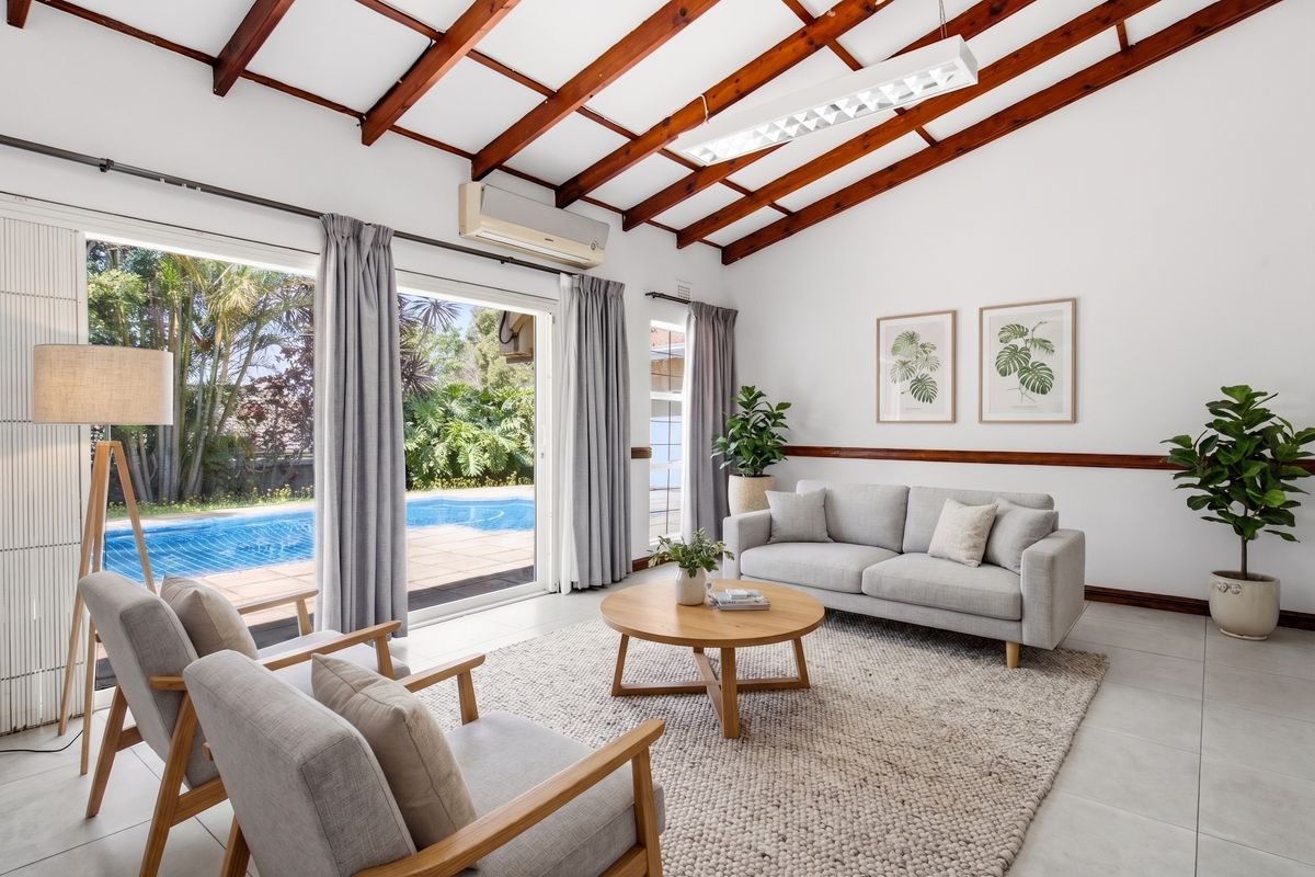 Bright living room featuring exposed timber ceiling beams, tiled flooring, and large sliding glass doors providing direct access to the pool area.