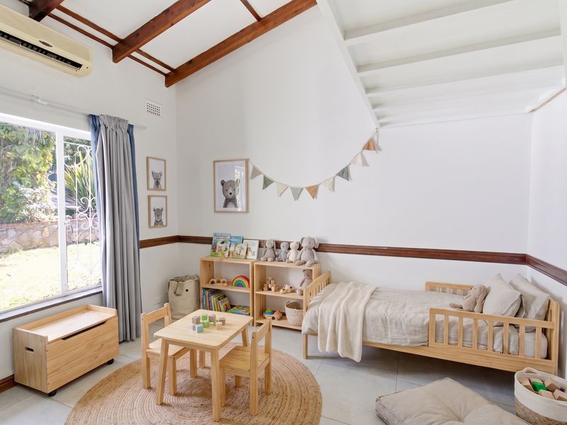 A bright bedroom featuring exposed timber ceiling beams, tiled flooring, a split-system air conditioner, and a large window overlooking the garden.
