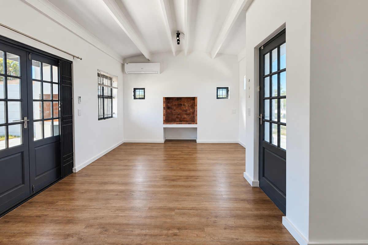A bright, empty living area featuring exposed beam ceilings, laminate flooring, and a built-in brick fireplace, with direct access to the exterior via french doors.