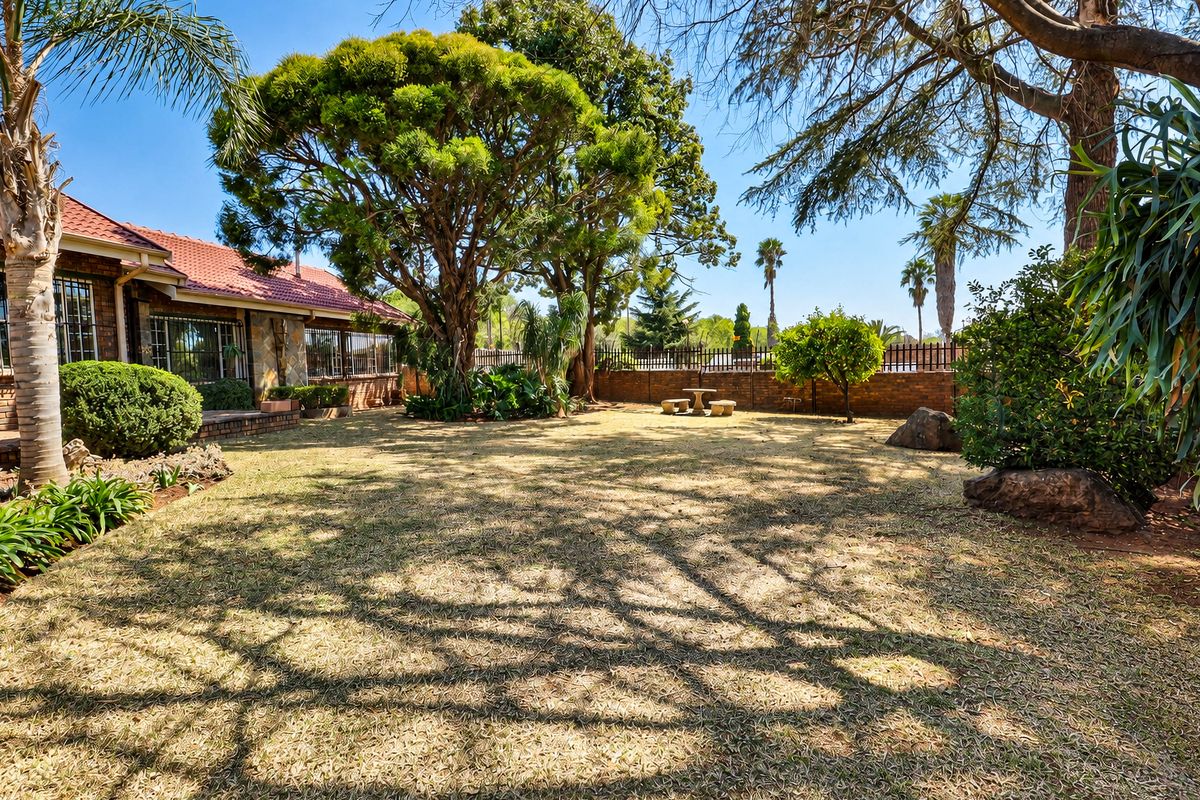 A spacious rear garden featuring mature trees, a brick boundary wall, and a view of the house with stone cladding and tiled roofing.
