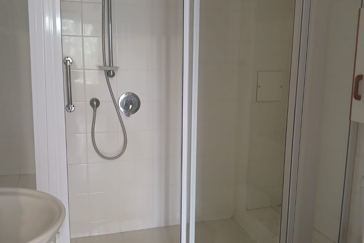 A functional bathroom featuring a shower enclosure with a handheld shower head, a pedestal basin, and a toilet, finished with white wall and floor tiles.