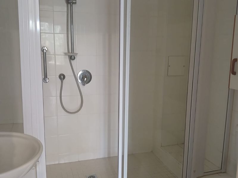 A functional bathroom featuring a shower enclosure with a handheld shower head, a pedestal basin, and a toilet, finished with white wall and floor tiles.