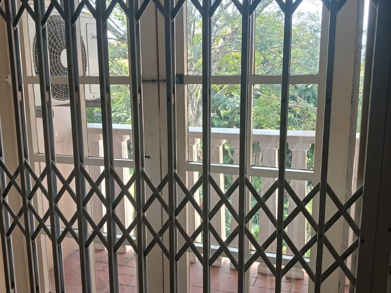 A view through a security gate onto a balcony featuring tiled flooring, a balustrade, and an external air conditioning unit.