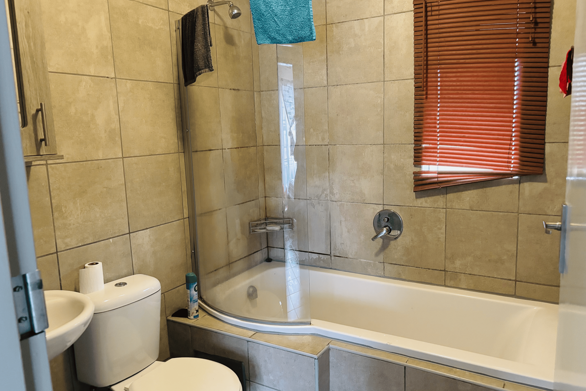 A functional bathroom featuring a combined bathtub and shower, pedestal basin, toilet, and neutral wall tiling with venetian blinds.