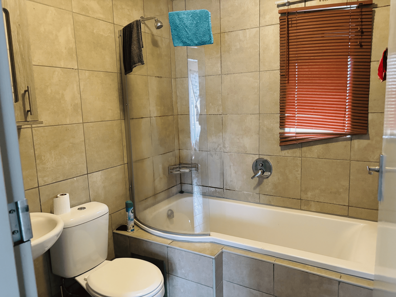 A functional bathroom featuring a combined bathtub and shower, pedestal basin, toilet, and neutral wall tiling with venetian blinds.