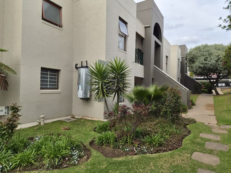 Rear exterior view of the townhouse complex featuring a landscaped garden, paved walkway, and external geyser unit.