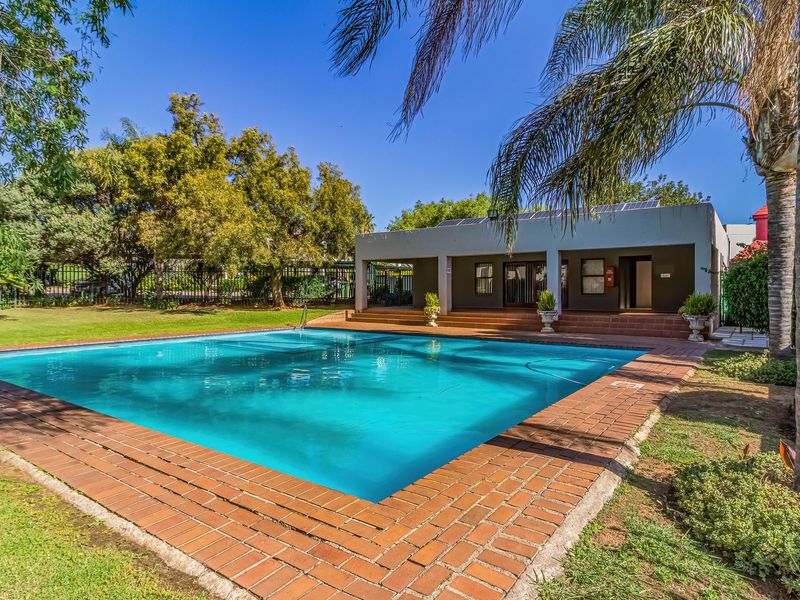 A well-maintained outdoor swimming pool area featuring brick coping, a spacious lawn, and an adjacent covered patio structure.