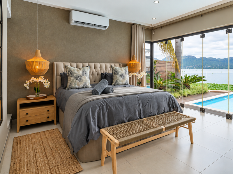 A modern, well-appointed bedroom featuring tiled floors, air conditioning, and floor-to-ceiling glass doors that open directly onto a private pool deck with water views.