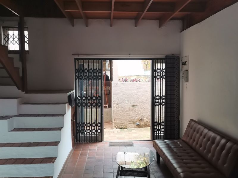 Living room featuring exposed timber beam ceilings, terracotta tile flooring, and a staircase, with direct access to an outdoor area through a security gate.