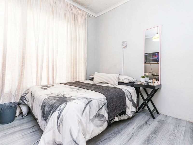 A compact bedroom featuring light-toned laminate flooring and large window curtains, showing signs of ceiling wear and needing cosmetic updates.
