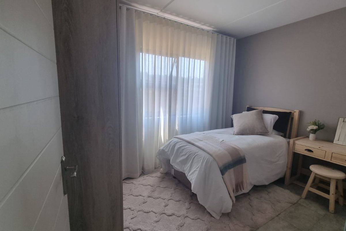 A well-lit bedroom featuring neutral wall tones, tiled flooring, and a large window with sheer curtains, providing a comfortable and functional living space.
