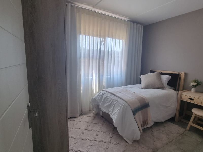 A well-lit bedroom featuring neutral wall tones, tiled flooring, and a large window with sheer curtains, providing a comfortable and functional living space.