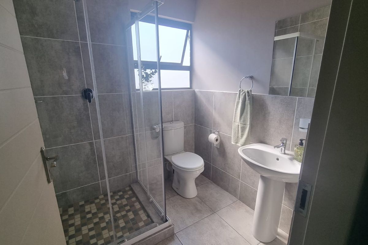 A modern bathroom featuring a pedestal basin, toilet, and a glass-enclosed shower with neutral grey wall and floor tiles.