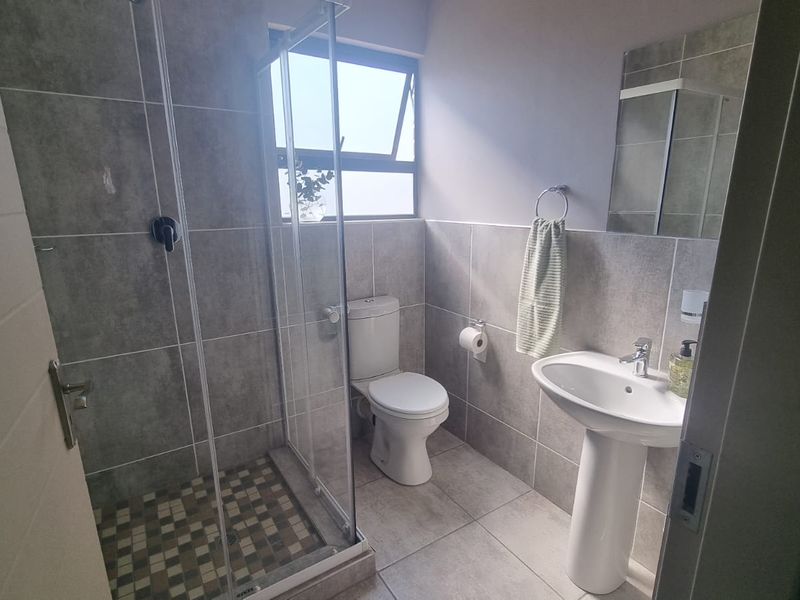 A modern bathroom featuring a pedestal basin, toilet, and a glass-enclosed shower with neutral grey wall and floor tiles.