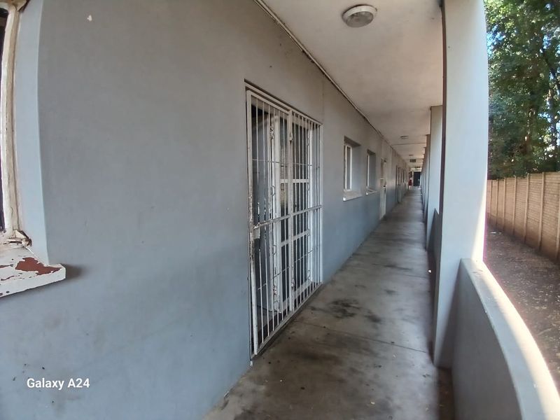 An exterior apartment building walkway featuring concrete flooring, security gates on unit entrances, and a parapet wall overlooking the grounds.