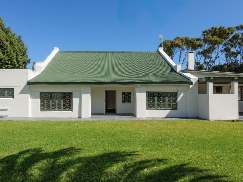 A rear view of a single-story house featuring a green corrugated metal roof, white exterior walls, security-barred windows, and a spacious lawn.