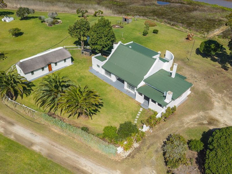 Aerial view of a rural property featuring a main house with a green metal roof, a separate thatched outbuilding, and expansive surrounding grounds.
