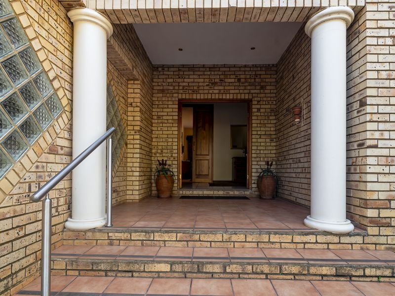 Covered front entrance featuring a brick facade, decorative columns, tiled steps with a stainless steel handrail, and a timber front door.