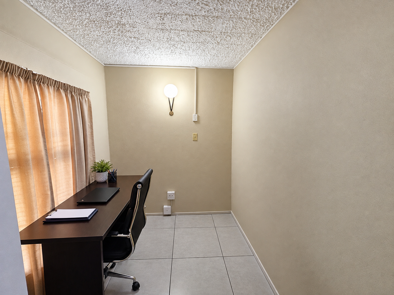 A compact office space featuring tiled flooring, a textured ceiling, and a wall-mounted light fixture with surface-mounted electrical conduit.