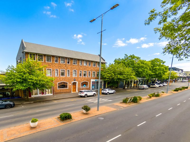 A street-level view of the apartment building featuring a classic brick facade, pitched roof, and established street-side landscaping.
