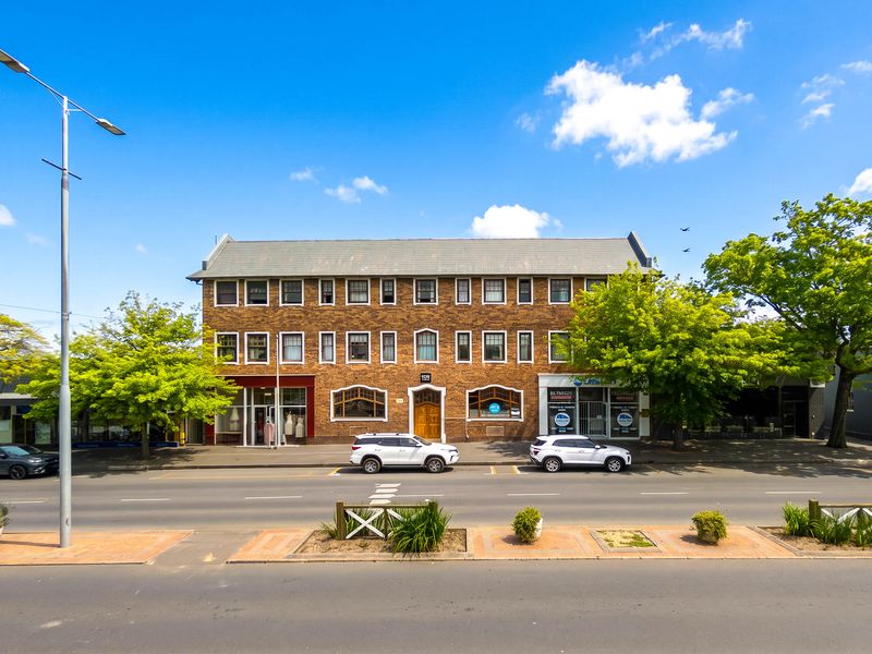 A three-story brick apartment building with ground-floor commercial storefronts, featuring a classic architectural design and pitched roof, situated along a street.
