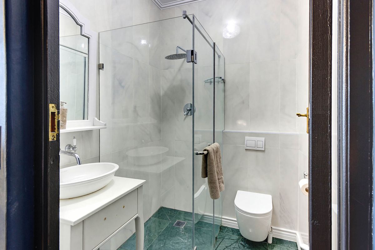 Modern bathroom featuring a glass-enclosed rain shower, wall-mounted toilet, vessel sink vanity, and elegant marble-style wall tiling with contrasting green marble flooring.