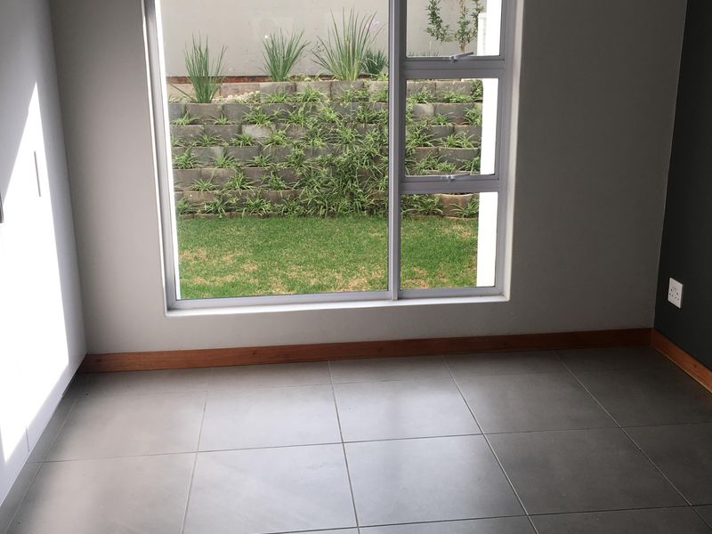 An empty bedroom featuring neutral grey floor tiles, timber skirting boards, and a large window overlooking a landscaped garden area.